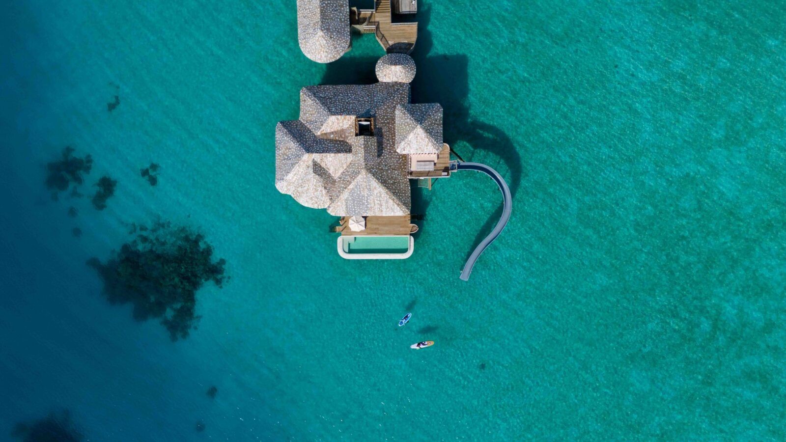 This image shows an aerial view of Crusoe villa at Soneva Secret Resort Maldives. With a swimming pool and water slide and a couple kayaking near villa.