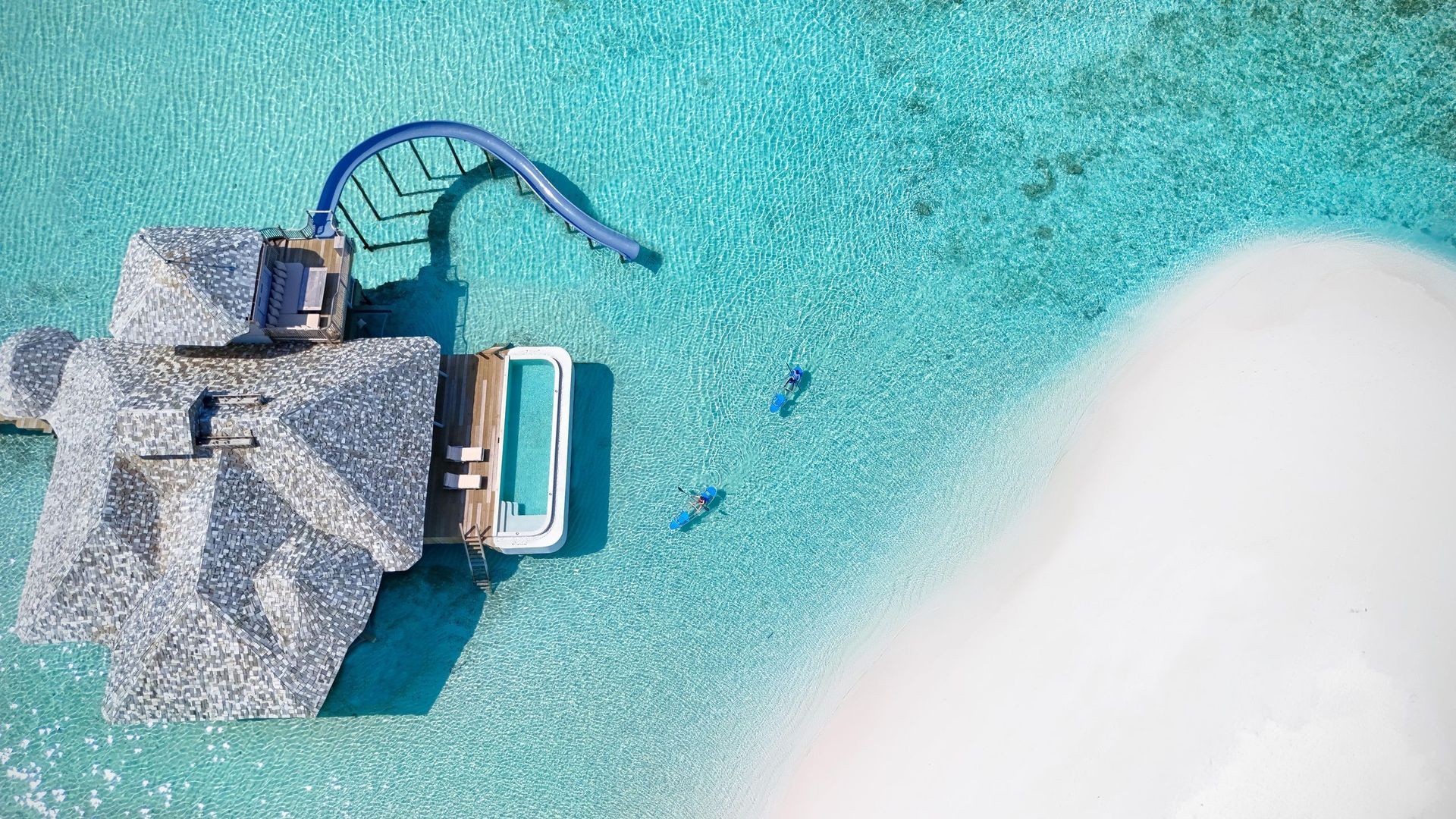 Aerial view of a resort featuring a large pool and a water slide, surrounded by lush greenery and sun loungers. 